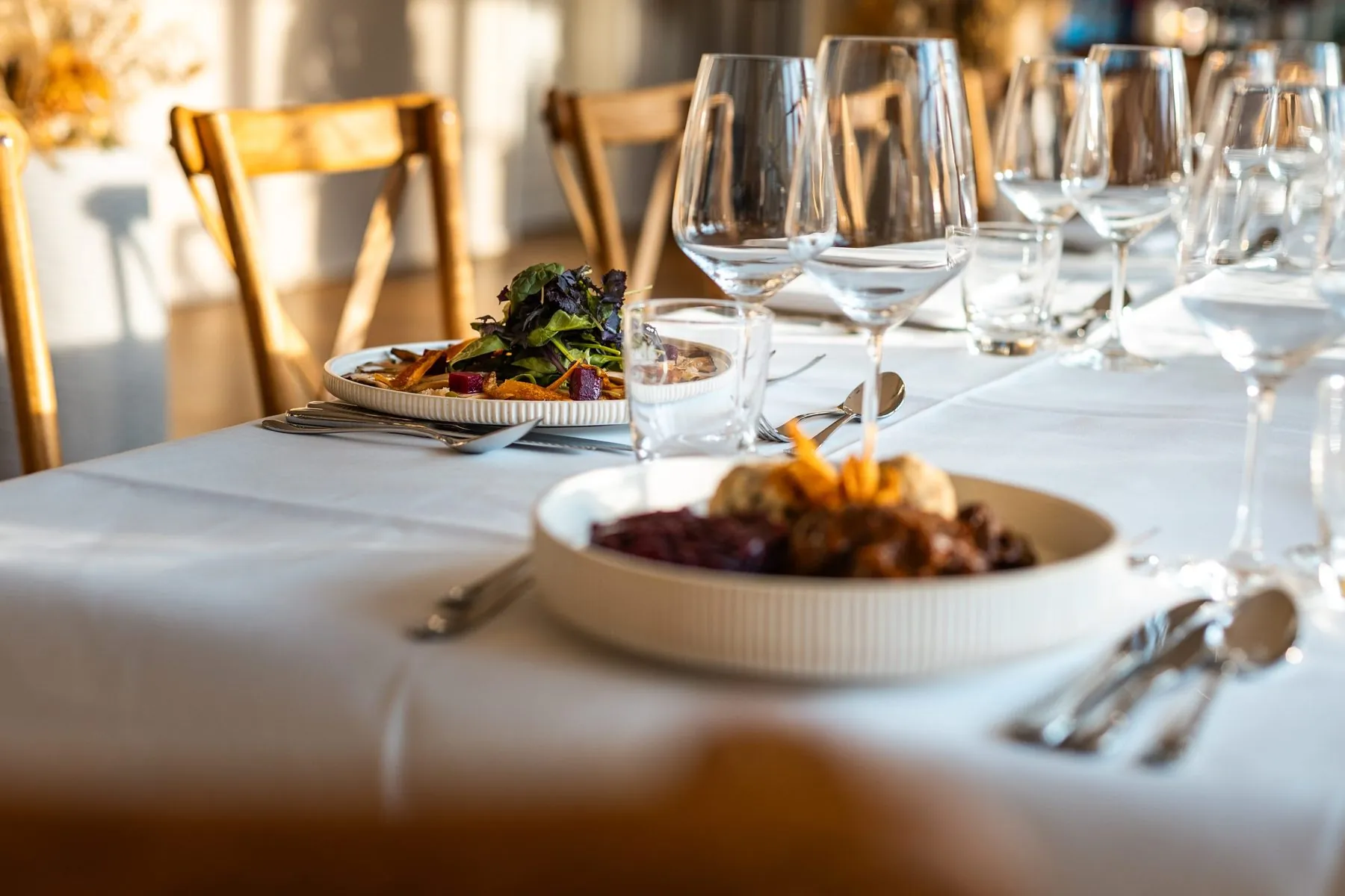 Weiß gedeckte lange Tafel mit Schmorgericht und Salat im warmen Seitenlicht