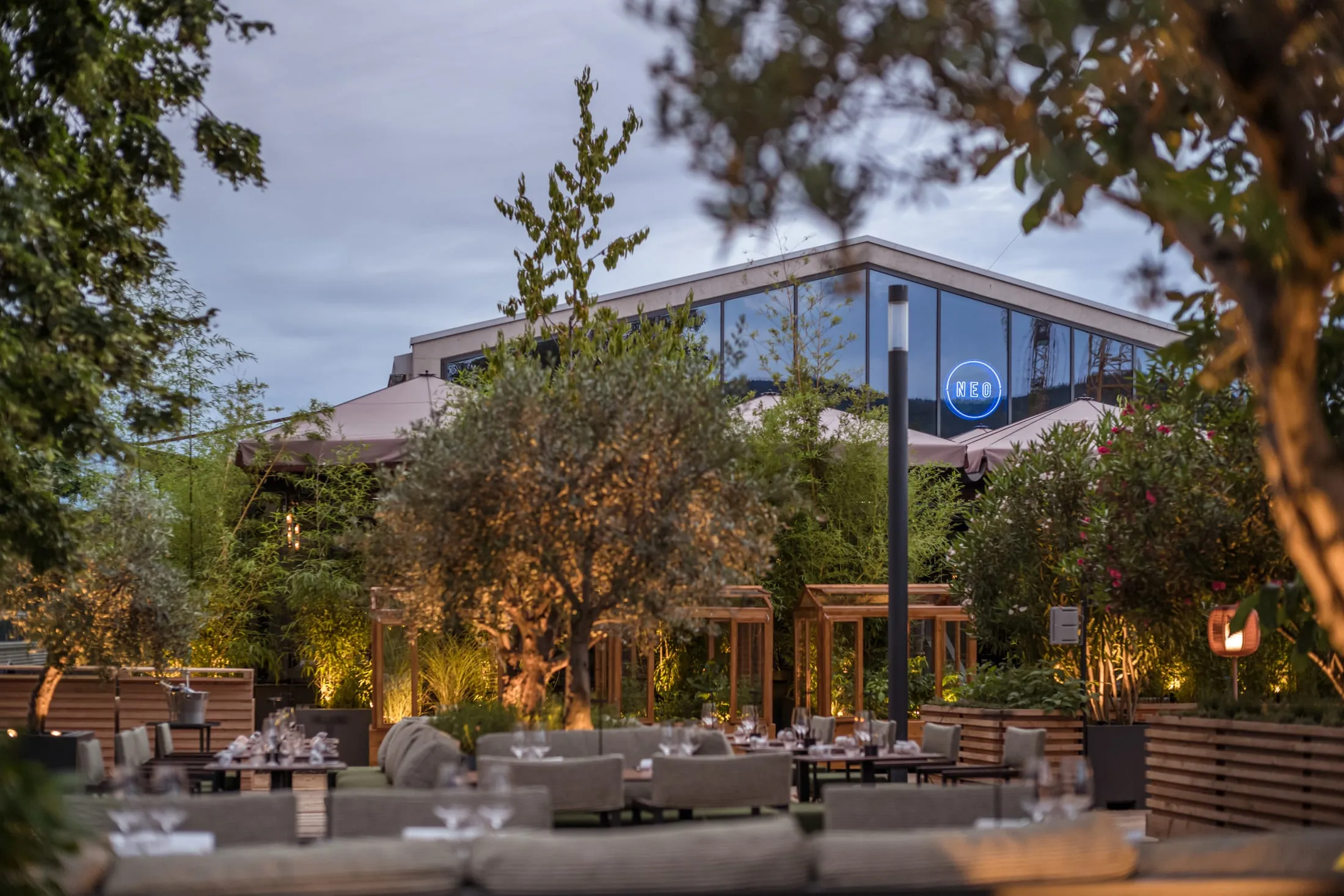 Garten des NEO in der Dämmerung mit angestrahltem Olivenbaum und gedeckter Lounge, dahinter die Glashalle
